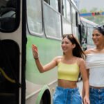 Two female friends smiling and waving to stop a public transportation bus on a sunny day in the city